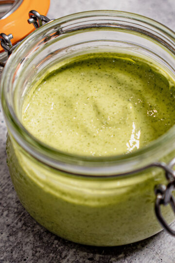 An open glass jar filled with creamy green sauce or dip, placed on a gray textured surface. The jar has a metal clasp and an orange rubber seal on the lid.
