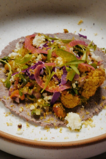 A blue corn tortilla topped with vegetables, crumbled cheese, and herbs, served on a white plate with a brown rim.