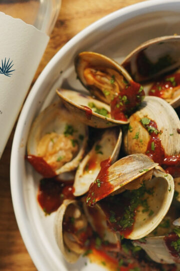 A white bowl of steamed clams topped with herbs and red sauce sits beside a partially visible bottle of Tres Agaves organic tequila on a wooden surface.