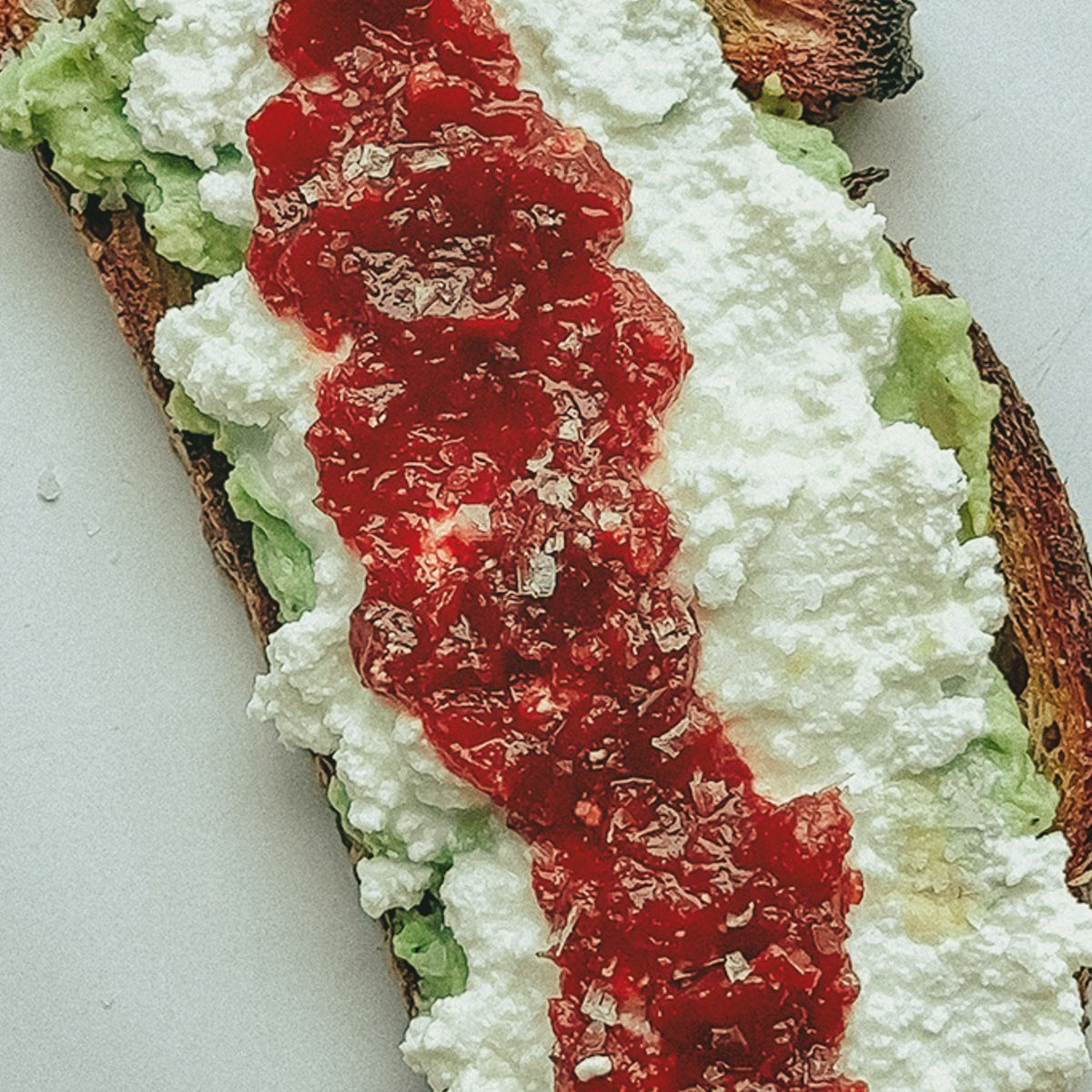 A slice of toasted bread topped with mashed avocado, ricotta cheese, and a stripe of red jam or preserve, served on a white surface.