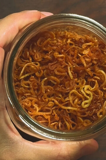 A hand holds a glass jar filled with crispy, golden-brown fried shallots, viewed from above on a wooden surface.