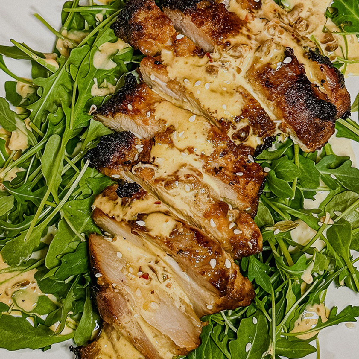 Grilled, sliced chicken breast with a creamy sauce served on a bed of fresh arugula leaves, garnished with sesame seeds.