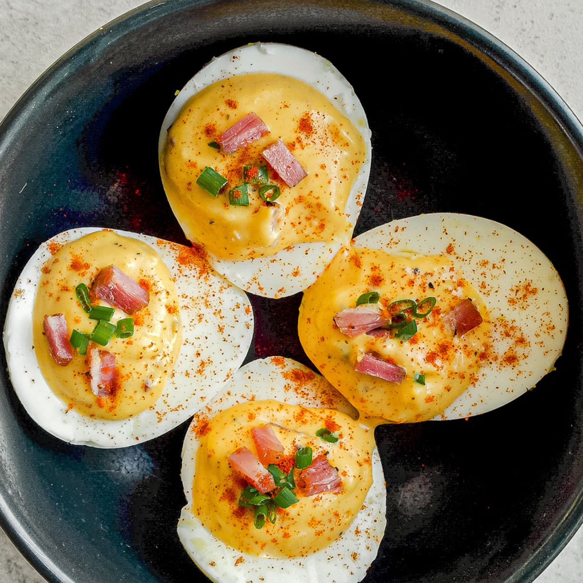 Four deviled egg halves topped with creamy yellow filling, diced bacon, chopped green onions, and a sprinkle of paprika, served in a black bowl on a light surface.
