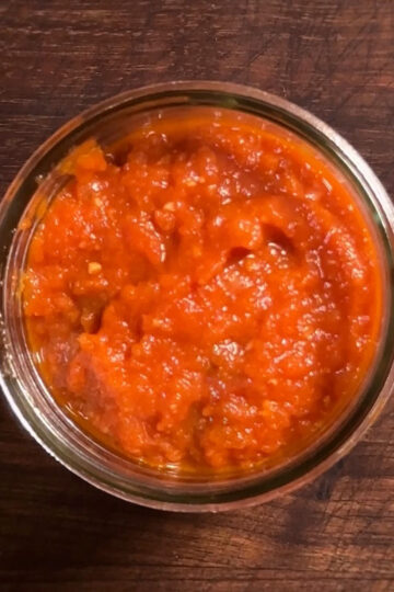 A small glass jar filled with bright red, chunky sauce sits on a dark wooden surface.