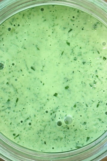 A close-up of a jar filled with creamy green dressing or sauce, speckled with chopped herbs, viewed from above on a wooden surface.