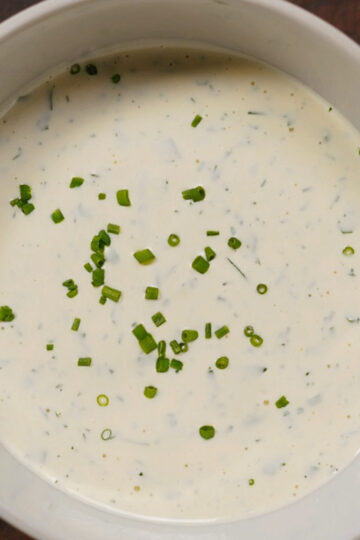 A white bowl filled with creamy ranch dressing, garnished with finely chopped green chives, sitting on a dark wooden surface.
