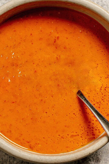 A bowl filled with bright orange, creamy sauce, with a metal spoon partially submerged inside. The bowl sits on a light gray textured surface.