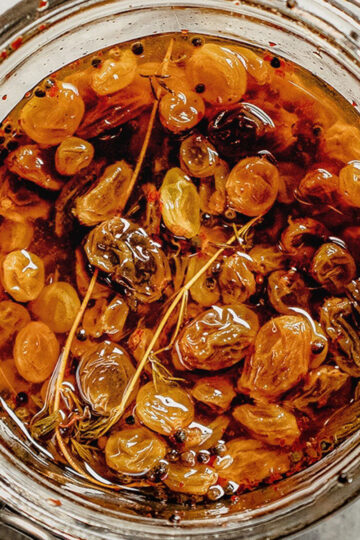 A close-up of a glass jar filled with golden raisins soaking in oil and spices, including visible mustard seeds and herbs. The jar is open, showing the rich, textured ingredients inside.
