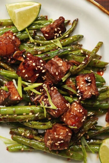 Stir-fried green beans and glazed beef cubes topped with sesame seeds and sliced green onions, served on a white plate with lime wedges.