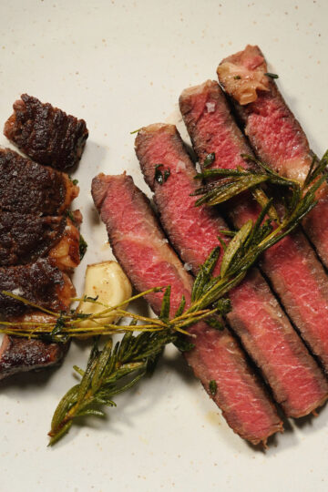 Slices of medium-rare steak arranged on a white plate, garnished with fresh herbs such as rosemary and thyme, and accompanied by a piece of roasted garlic.