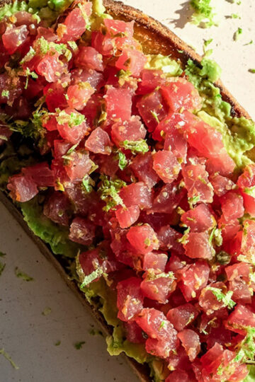 A slice of toasted bread topped with mashed avocado and diced raw tuna, garnished with lime zest. Some lime zest is scattered on the plate around the toast.