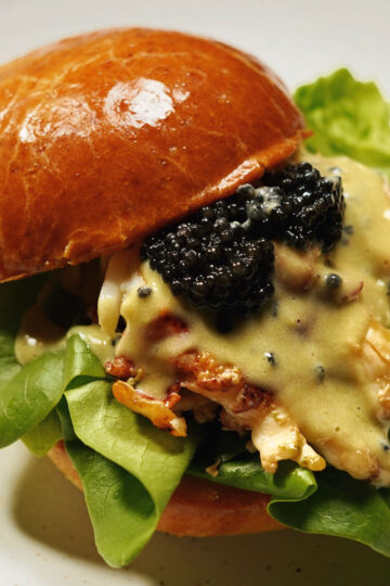 A gourmet sandwich with a glossy brioche bun, fresh green lettuce, lobster meat, creamy sauce, and a generous topping of black caviar.