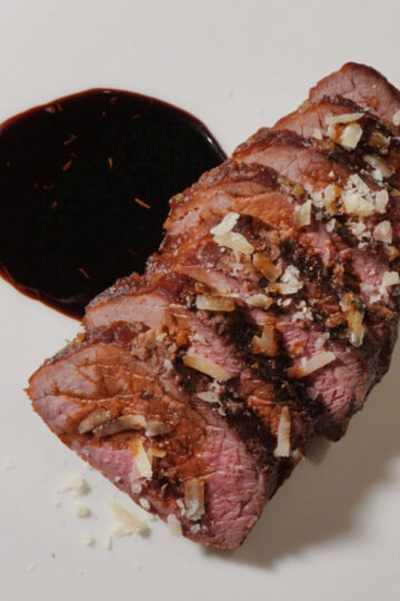 Five slices of medium-rare steak topped with grated cheese are arranged in a fan shape on a white plate, next to a pool of dark sauce.