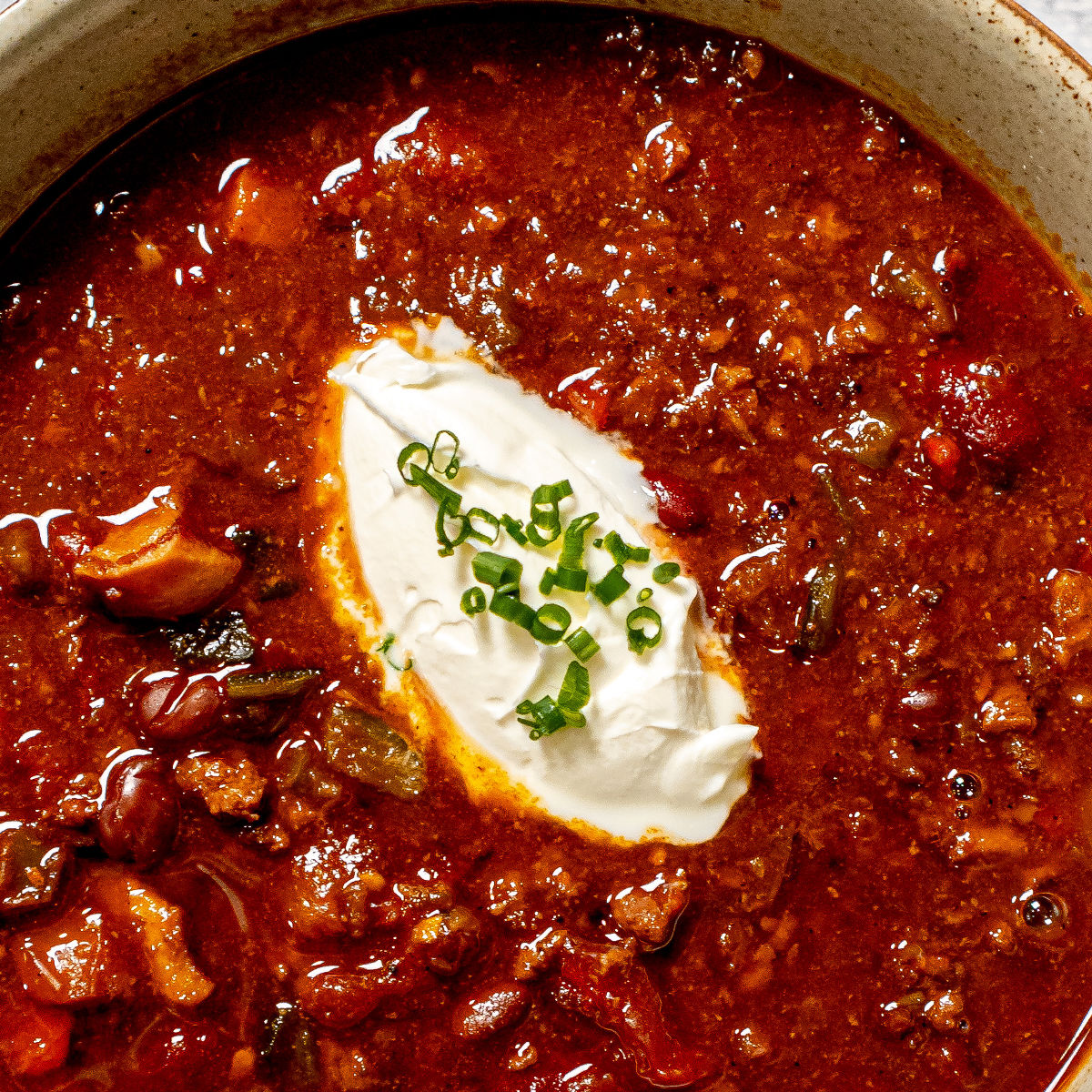 Close-up of a bowl of chili topped with a dollop of sour cream and chopped green onions, showing a rich, thick, reddish-brown stew with visible chunks of vegetables and meat.