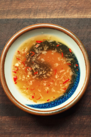 A small ceramic bowl of Vietnamese dipping sauce with chopped red chili, garlic, and floating black and orange liquids, placed on a wooden surface.