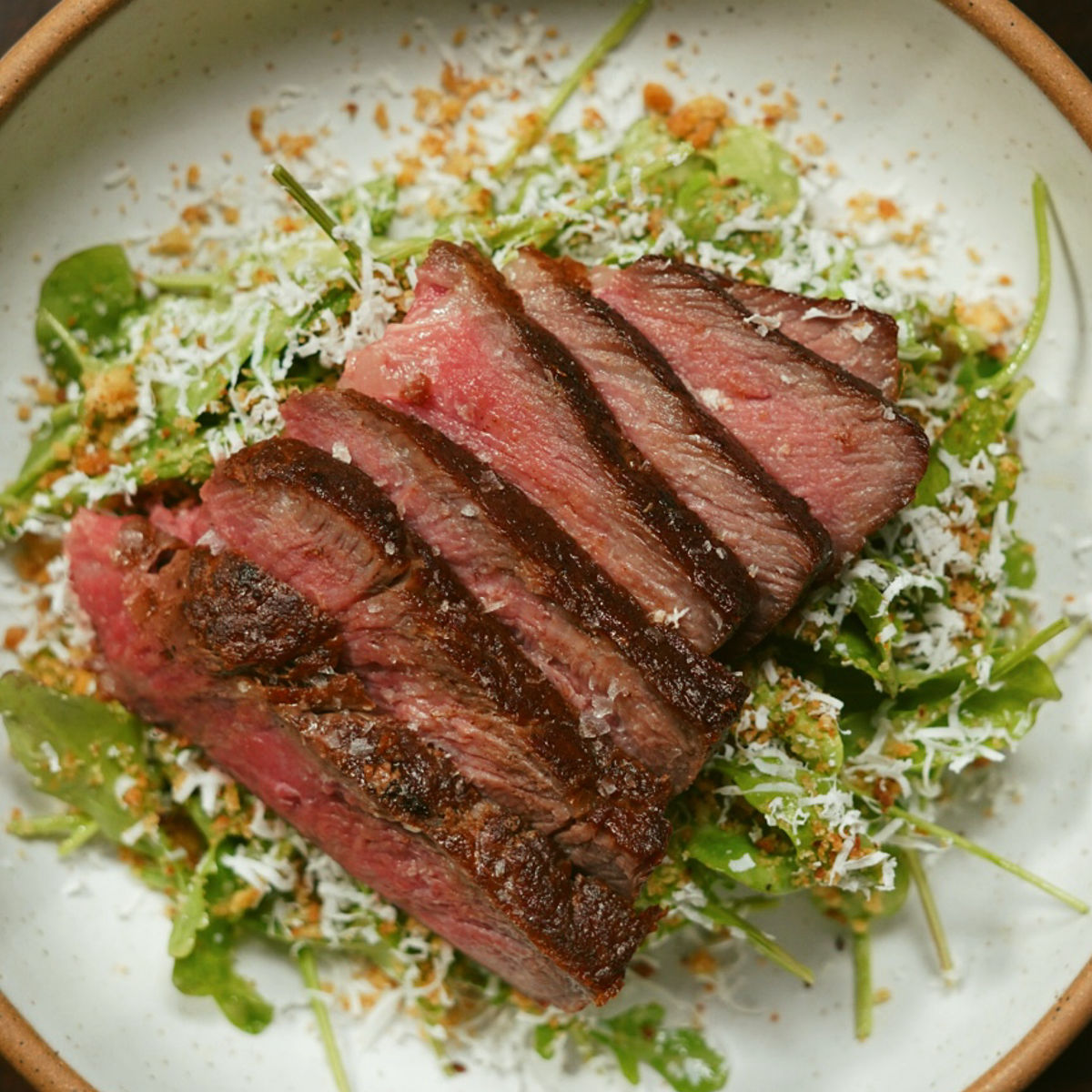 A bowl of sliced medium-rare steak served on a bed of fresh greens, topped with grated cheese and crunchy breadcrumbs.