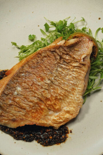 A cooked fish fillet with crispy golden-brown skin rests on a bed of fresh green herbs and is served with a dark, chunky sauce on a beige plate.