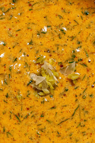 A close-up of a creamy, orange-yellow sauce speckled with green herbs and spices, topped with a small amount of finely chopped pickles or vegetables in the center.