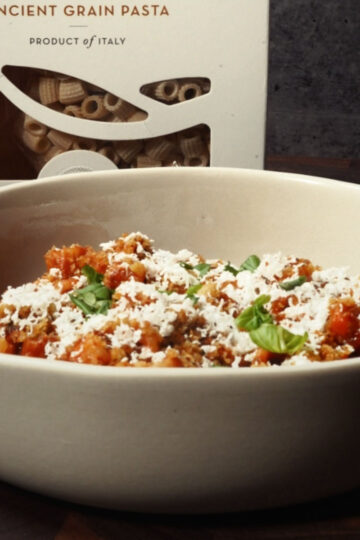 A bowl of pasta with tomato sauce, topped with grated cheese and fresh basil leaves, sits in front of a box labeled Ancient Grain Pasta. The pasta box is partially open, revealing tube-shaped noodles inside.