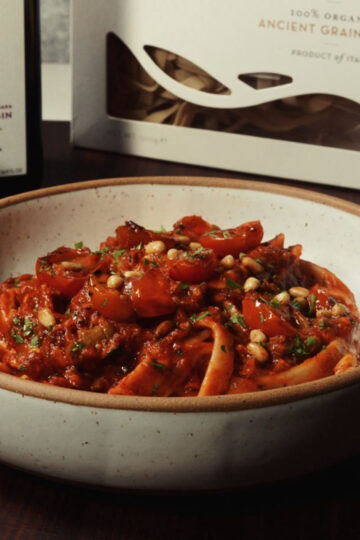 A bowl of pasta topped with tomato sauce, cherry tomatoes, herbs, and pine nuts sits on a dark table. In the background are a bottle of extra virgin olive oil and a box labeled Ancient Grain Pasta.