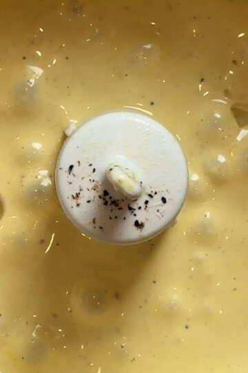 Close-up of creamy yellow sauce with black pepper specks in a food processor bowl, with the white blade attachment visible in the center.