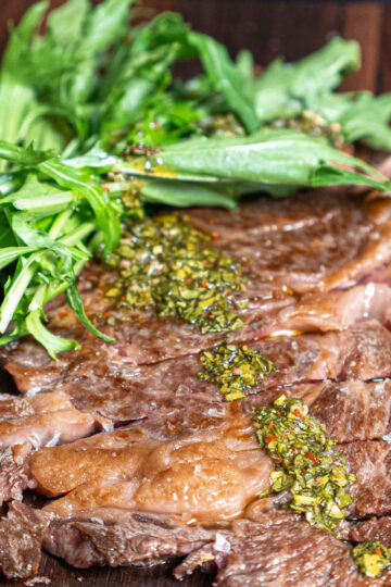 Sliced grilled steak topped with chimichurri sauce, served on a wooden board with a side of fresh green leafy salad.