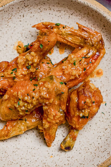 A speckled beige bowl filled with several glazed chicken wings, garnished with chopped herbs and minced garlic.