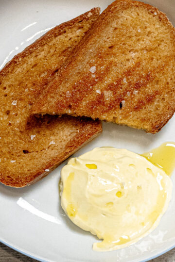 Two slices of toasted brown bread rest on a white plate next to a serving of creamy butter topped with a drizzle of olive oil.