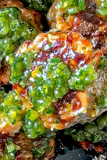 Close-up of grilled meatballs topped with a vibrant green chimichurri sauce, giving the dish a fresh and appetizing appearance. The meatballs are charred in some areas, adding texture and color contrast.