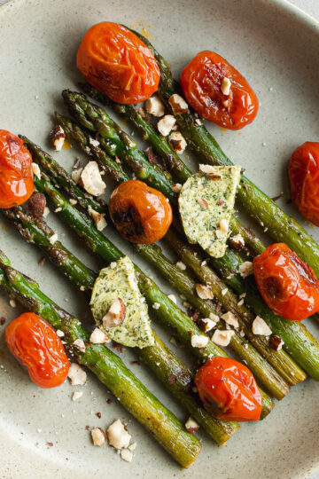 A beige plate with roasted asparagus spears, blistered cherry tomatoes, dollops of herbed butter, and chopped nuts sprinkled on top.