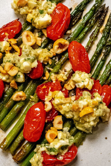 Grilled asparagus spears topped with roasted cherry tomatoes, chopped nuts, and a creamy herbed cheese mixture on a white plate.