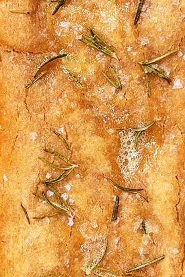 A close-up of a rectangular piece of focaccia bread topped with coarse salt and rosemary sprigs on a light-colored surface.
