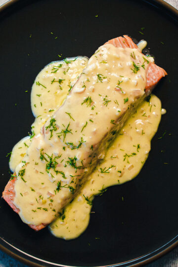 A fillet of salmon on a black plate, topped with creamy yellow dill sauce and garnished with fresh chopped dill.