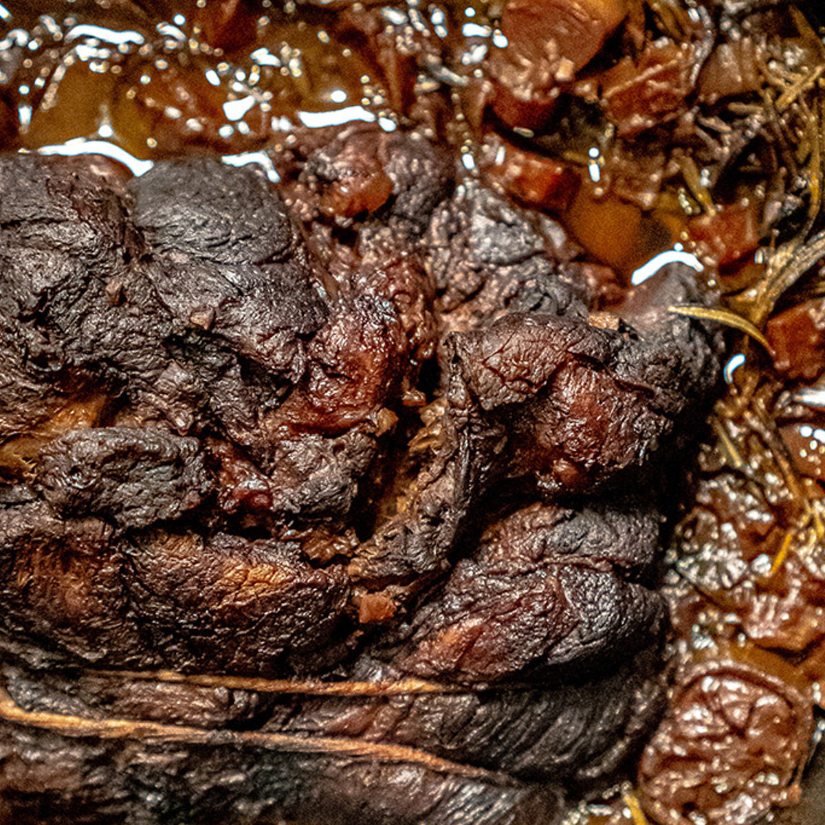 Close-up of a cooked pot roast with a dark, seared exterior, surrounded by rich, brown juices and pieces of vegetables. The meat appears tender and moist, sitting in a flavorful sauce.