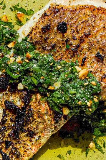 Close-up of seasoned, grilled fish fillet topped with a fresh green herb sauce containing chopped parsley, pine nuts, and garlic, all on a plate with a drizzle of olive oil.