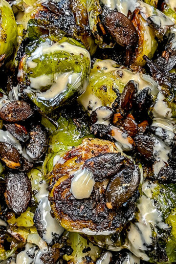 Close-up of roasted Brussels sprouts with crispy, charred edges, drizzled with a creamy, light-colored sauce. The sprouts are caramelized and slightly browned, highlighting their texture and glossy finish.