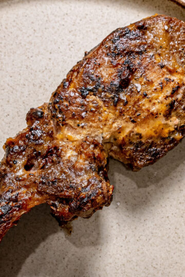 A cooked, seasoned lamb chop with a bone is placed on a beige plate. The meat has a slightly charred, crispy exterior and visible spices.