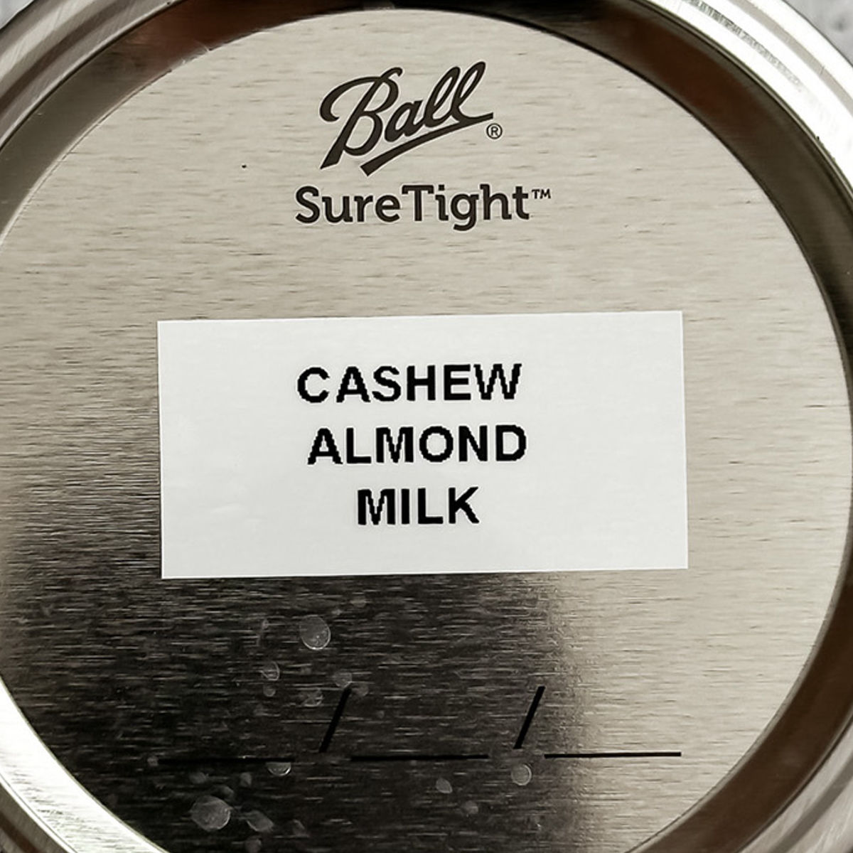 A jar lid labeled Ball SureTight with a white sticker that reads CASHEW ALMOND MILK in bold black letters.