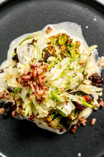 A gourmet salad with shaved greens, bits of crispy bacon, and roasted vegetables, served on a creamy white sauce, arranged artfully on a black plate.