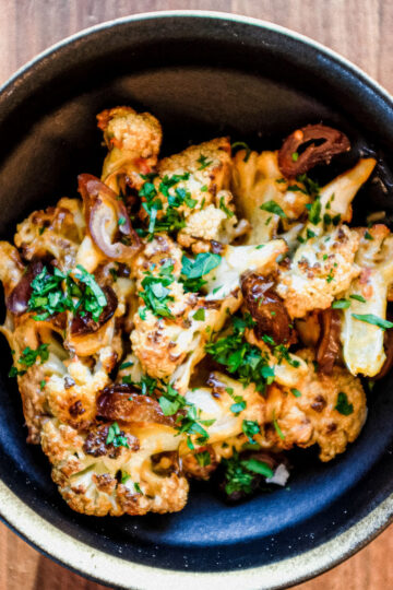 A black bowl filled with roasted cauliflower, melted cheese, mushrooms, and chopped herbs, placed on a wooden table.