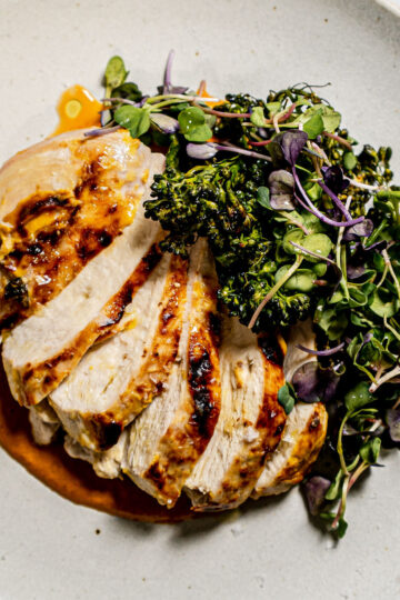 Sliced grilled chicken breast with sauce, served with fresh greens and broccolini on a white plate.
