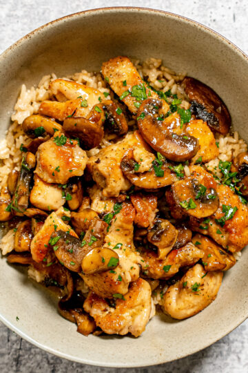 A bowl of brown rice topped with sautéed chicken pieces, sliced mushrooms, and chopped parsley, all coated in a savory sauce.