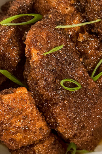 Close-up of crispy, golden-brown fried chicken pieces garnished with sliced green onions, served in a bowl with a bit of sauce at the bottom.