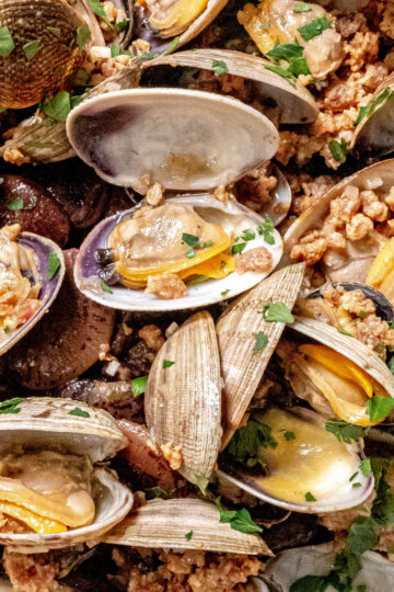 A close-up of cooked clams in open shells, topped with chopped parsley and crumbled sausage, showcasing a seafood dish with vibrant colors and varied textures.