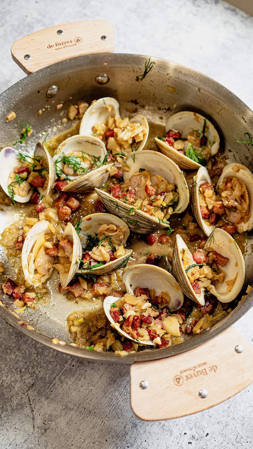A pan filled with cooked clams, garnished with chopped herbs and diced bacon or pancetta, sits on a gray countertop. The dish is saucy and aromatic, with pieces of onion or garlic visible among the clams.
