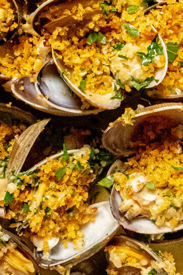 Close-up of baked clams topped with golden breadcrumbs and chopped parsley, showing open shells filled with savory stuffing and herbs.