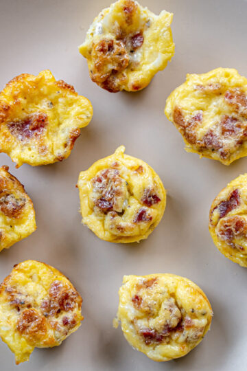 Eight mini egg muffins with visible bits of bacon and cheese are arranged on a round beige plate. The muffins are golden brown on top and appear fluffy and baked.