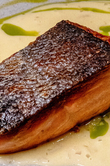 A close-up of a seared salmon fillet with crispy skin, served on a creamy white sauce and drizzled with green oil, presented on a white plate.