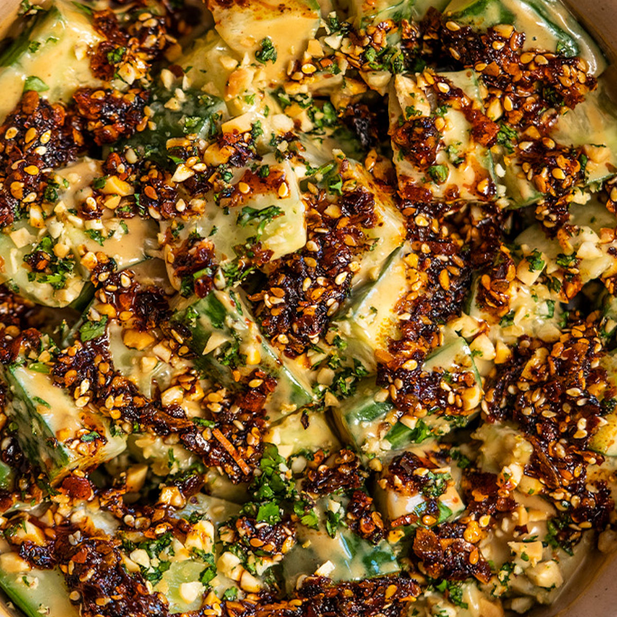 Close-up of sliced cucumbers coated in a creamy, spicy chili oil sauce with sesame seeds and chopped herbs, creating a vibrant, textured salad with visible seasonings and fresh ingredients.