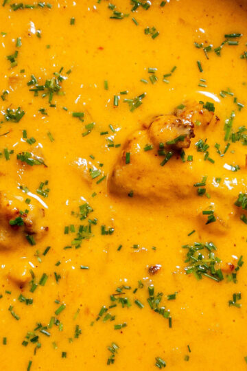 Close-up of a creamy, orange soup with visible chunks, garnished with chopped green herbs sprinkled on top. The surface is glossy, suggesting a rich, smooth texture.